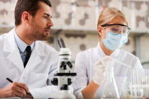 Two people in lab coats and masks looking at a microscope.