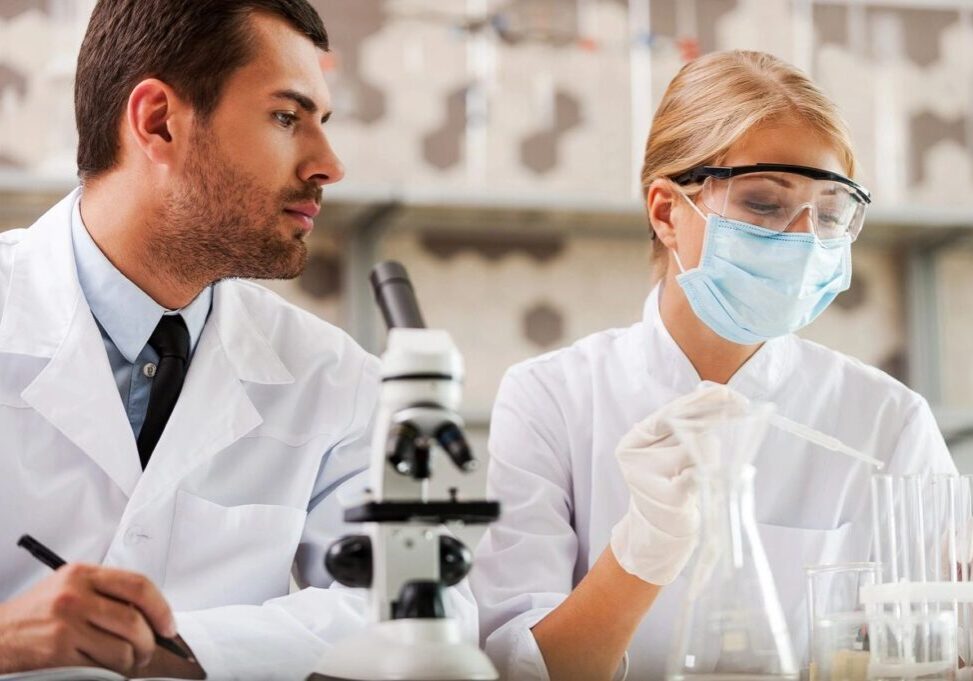 Two people in lab coats and masks looking at a microscope.